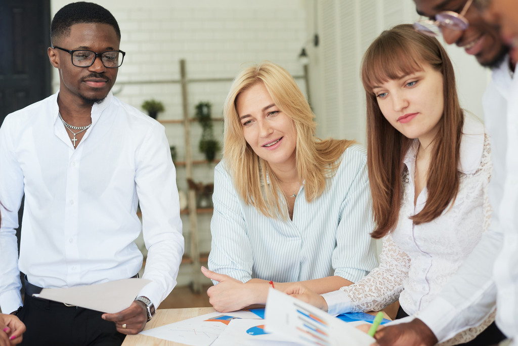 Neljä henkilöä pöydän ympärillä keskustelemassa ja tarkastelemassa asiakirjoja tai kaavioita toimistoympäristössä, taustalla tiiliseinä ja hyllyjä kasveilla.