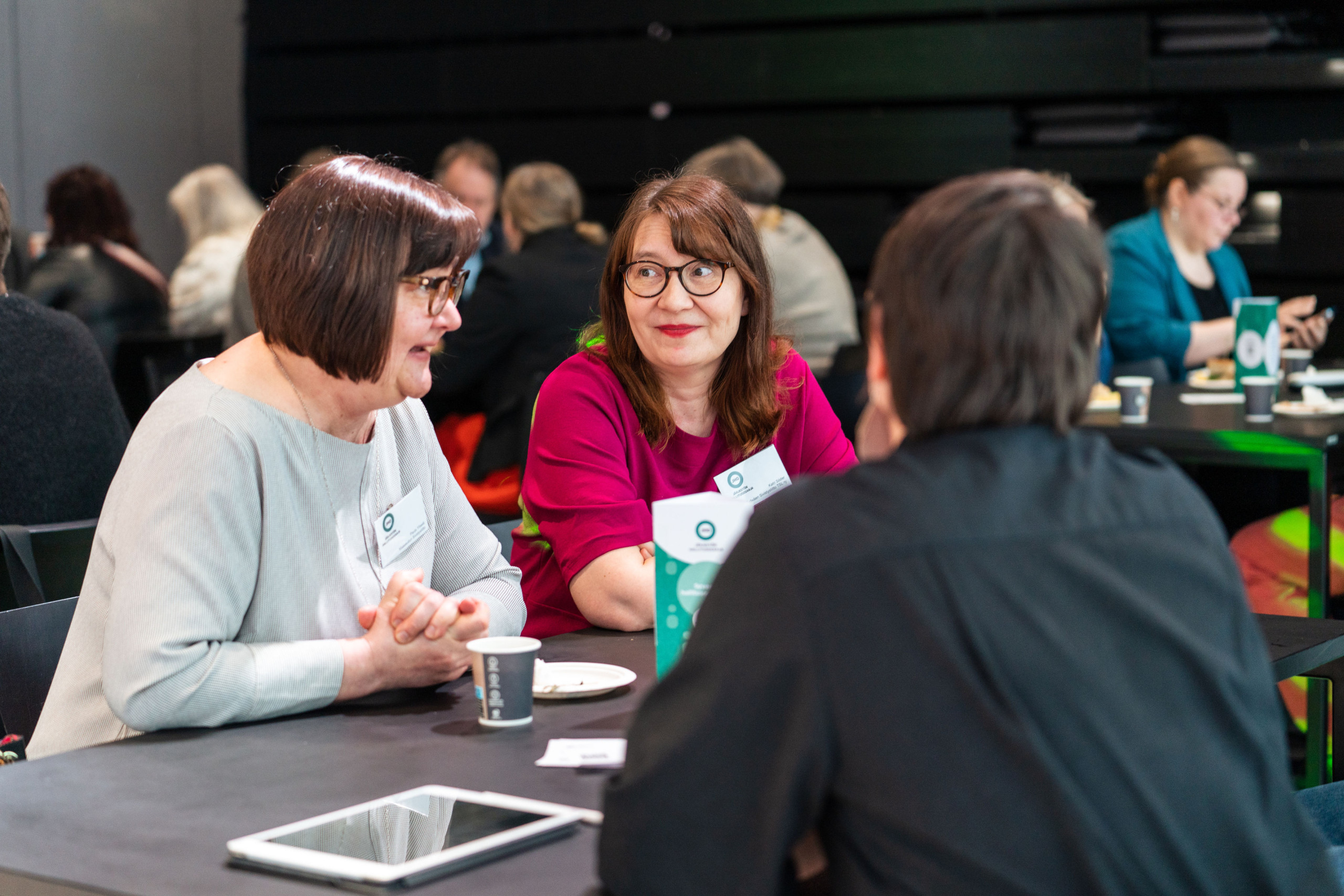 Kolme henkilöä keskustelemassa pöydän ääressä tapahtumassa, jossa taustalla näkyy muita osallistujia. Pöydällä on kahvi ja älytabletti ja muistiinpanoja.